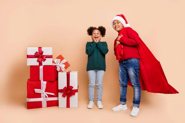 2人の子供の写真は新年を祝う男の子は袋の贈り物を運ぶx-mas帽子ジャンパー孤立ベージュ色の背景を身に着けている — ストック写真