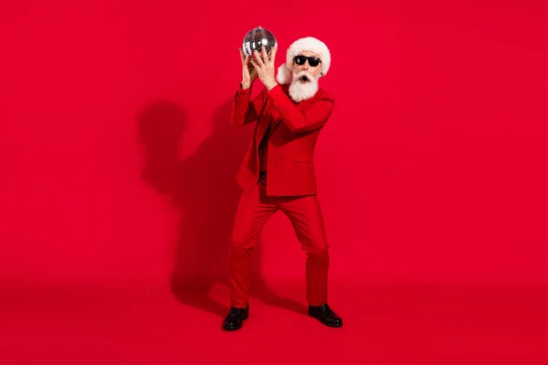 Komik gri saçlı yaşlı adamın tam vücut fotoğrafı top tutarken kırmızı takım elbise giyip kırmızı arka planda izole edilmiş Noel Baba şapkası giyiyor. — Stok fotoğraf