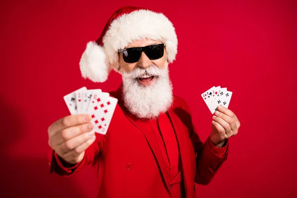 Foto di allegro eccitato Babbo Natale tenere le carte consiglio dei fan gioco d'azzardo indossare occhiali da sole x-mas cappello vestito su sfondo di colore rosso — Foto Stock