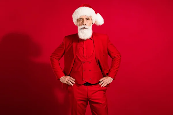 Photo of serious gentleman old man hold hands waist santa claus hat suit isolated on red color background