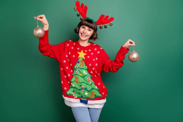 Photo of pretty sweet young woman dressed red pullover antlers smiling holding new year balls isolated green color background — Stock Photo, Image