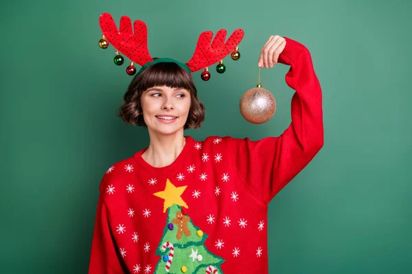 Фото чарівної красивої молодої жінки, одягнені в червоний пуловер, посміхаються, дивлячись, що тримає новорічний м'яч ізольований зелений колір фону — стокове фото