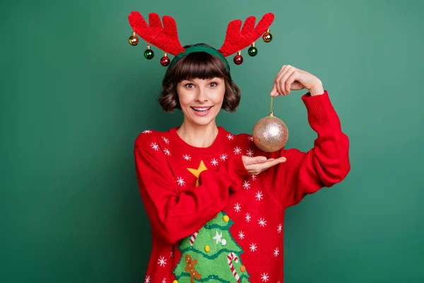Фото смішних схвильованих жіночого одягу светр роги, що тримають різдвяну прикрасу ялинки, усміхнений ізольований зелений колір фону — стокове фото