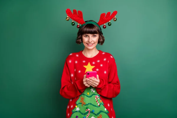 Photo of funky cute young woman dressed red pullover antlers smiling typing modern device isolated green color background — Stock Photo, Image