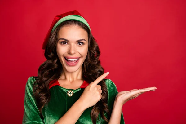 Foto di dolce eccitato giovane donna vestito elfo costume sorridente braccio che tiene punta vuoto spazio isolato colore rosso sfondo — Foto Stock