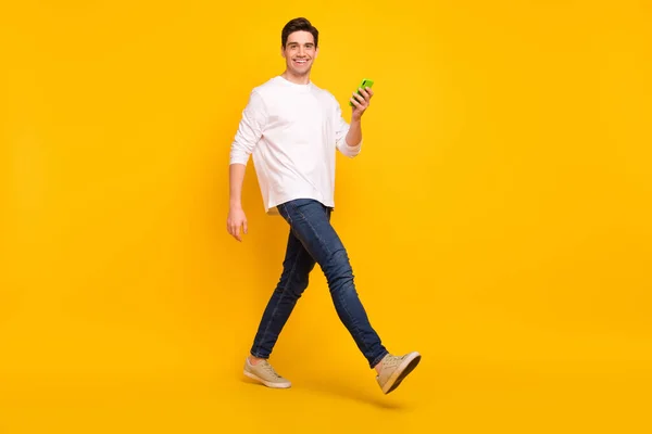 Foto de longitud completa de divertido morena millennial chico ir mantenga teléfono desgaste camisa jeans zapatillas aisladas sobre fondo amarillo — Foto de Stock
