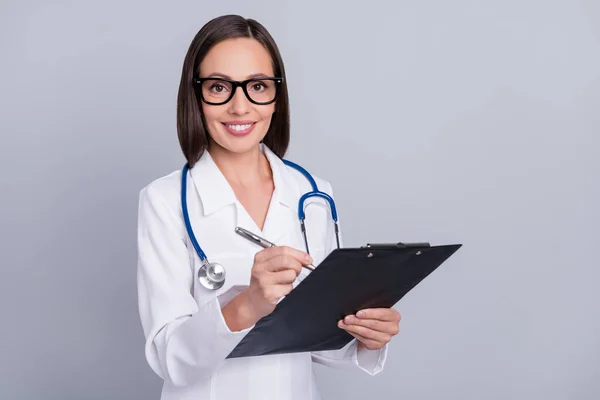 Φωτογραφία του γλυκού αστείο γιατρό κυρία φορούν λευκό επίσημη γυαλιά βραχίονα χαμογελώντας γράψιμο πρόχειρο απομονωμένο γκρι φόντο χρώμα — Φωτογραφία Αρχείου