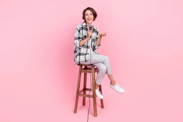 Plná délka fotografie mladé ženy šťastný pozitivní sedět stolička mluvit mikrofon výkon izolované přes růžové barvy pozadí — Stock fotografie