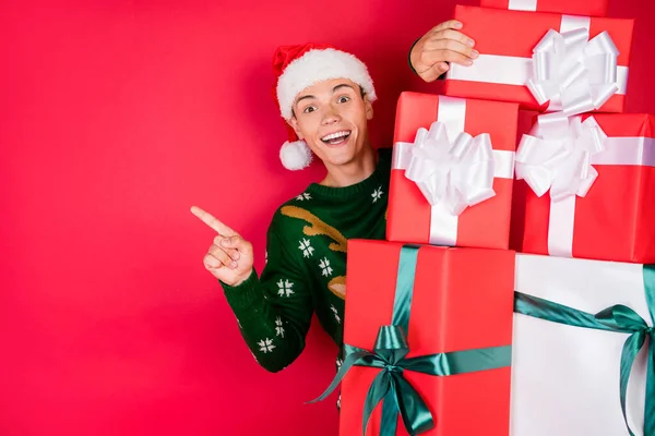 Фотопортрет человек улыбающийся парень в праздничной шляпе свитер прячется за подарки указывая копирайс изолированные ярко-красный цвет фона — стоковое фото