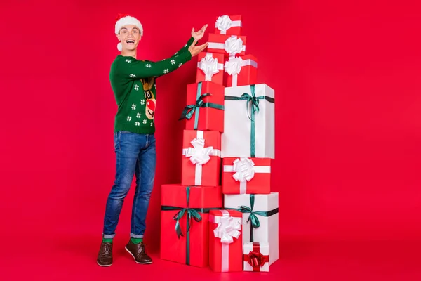 Ganzkörpergröße Foto überglücklicher Kerl trägt festliche Mütze zeigt Geschenk auf Weihnachten isoliert lebendige rote Farbe Hintergrund — Stockfoto