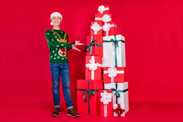 Ganzkörpergröße Foto glücklicher Kerl trägt festliche Mütze zeigt Geschenk-Boxen isoliert lebendige rote Farbe Hintergrund — Stockfoto