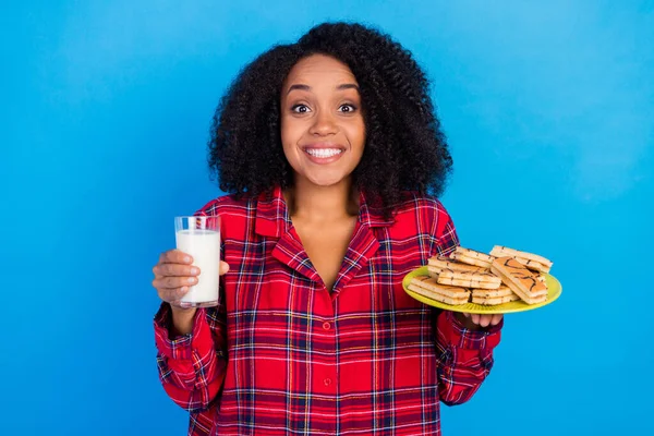 楽観的なミレニアムの写真ブルネットの女性はお菓子を食べるミルクは、青の色の背景に隔離された赤いチェッカーのスリーパーウェアを着用 — ストック写真