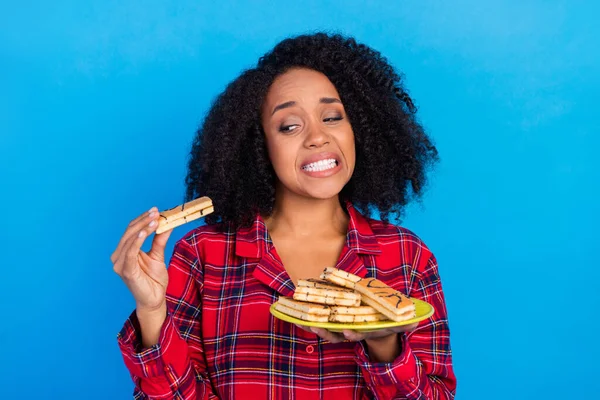 悲しい若いブルネットの女性の写真不健康な食べ物を食べる赤いチェックされたスリーパーの着用青の色の背景に隔離された — ストック写真