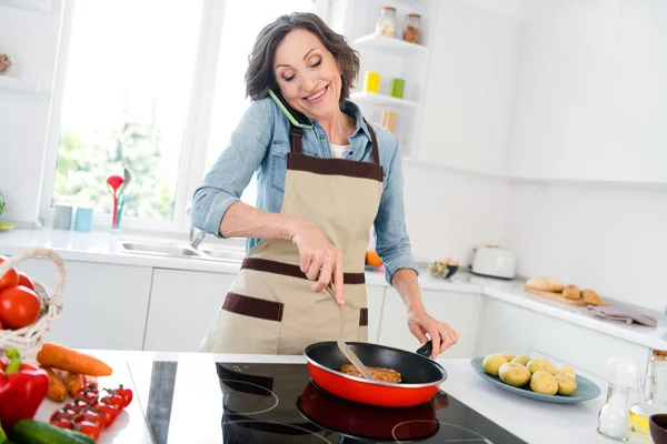 Foto av mogen rolig dam kock kött prata telefon bära förkläde jeans skjorta i köket ensam — Stockfoto