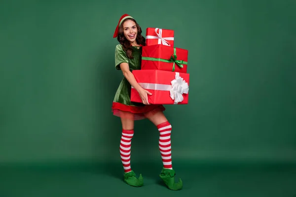 Full längd kroppsstorlek syn på ganska glad flicka nisse föra tunga gåvor isolerade över grön färg bakgrund — Stockfoto
