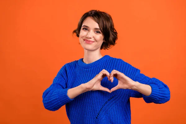 Foto van flirterige brunette millennial dame tonen hart slijtage blauwe trui geïsoleerd op oranje kleur achtergrond — Stockfoto