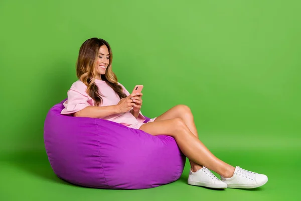 Foto de comprimento total da mulher feliz positiva sentar cadeira segurar as mãos telefone sorriso isolado no fundo cor verde — Fotografia de Stock