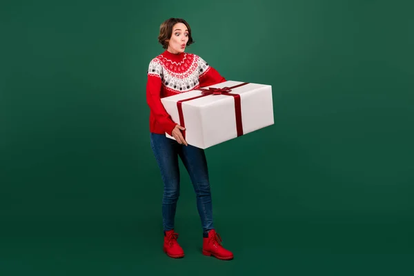 Longitud completa vista del tamaño del cuerpo de la atractiva chica sorprendida sosteniendo llevando pesada caja de regalo grande aislado sobre fondo de color verde — Foto de Stock