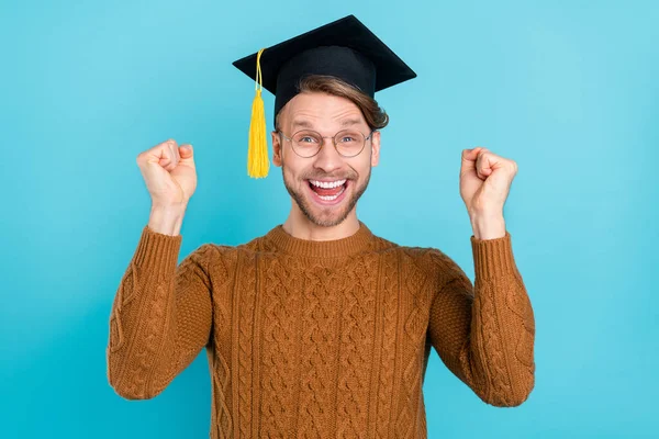 Foto van zoete charmante jonge heer dragen bruine pullover bachelor pet bril rijzende vuisten glimlachen geïsoleerde blauwe kleur achtergrond — Stockfoto