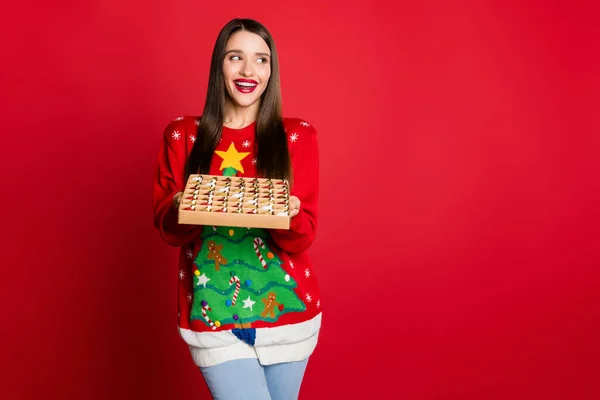 Фото шокованої солодкої молодої леді носити прикрашений светр, що тримає іграшкову коробку, дивлячись на порожній простір ізольований фон червоного кольору — стокове фото