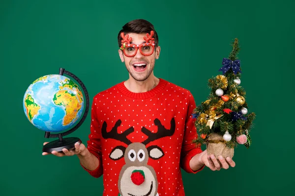 Porträt von attraktiven trendigen fröhlichen Kerl mit kleinen Baum Globus Spaß Winter isoliert über grüne Farbe Hintergrund — Stockfoto