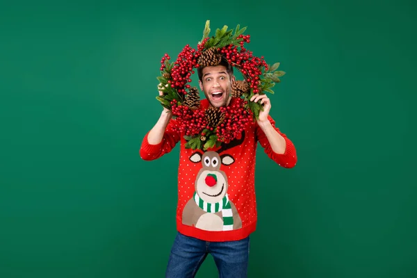 Foto Porträt Mann trägt lustigen Pullover hängen Weihnachtskranz starrt verrückt isoliert grüne Farbe Hintergrund — Stockfoto