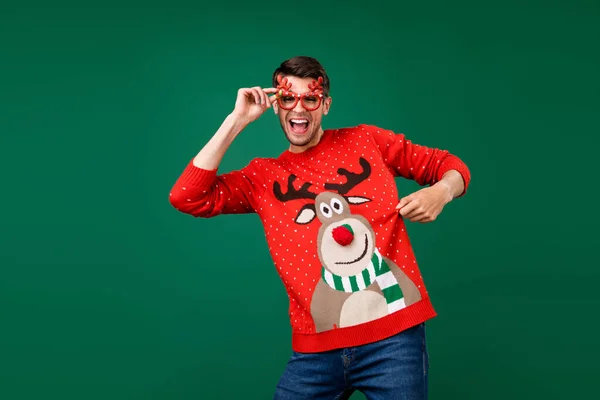 Мужчина в смешных очках танцует на рождественской вечеринке, показывая уродливый свитер изолированного зеленого цвета фона — стоковое фото