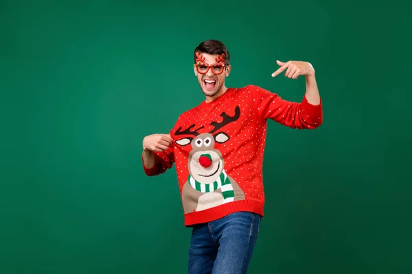 Foto do jovem feliz sorriso positivo ponto dedo decoração rena jumper óculos traje isolado sobre cor verde fundo — Fotografia de Stock