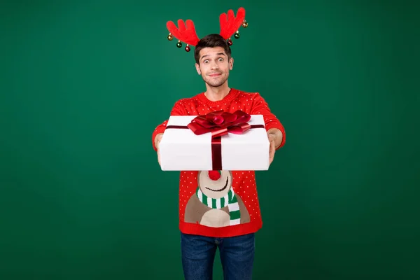 Photo de jeune homme heureux sourire positif tenir boîte cadeau donner partie porter bandeau renne isolé sur fond de couleur verte — Photo