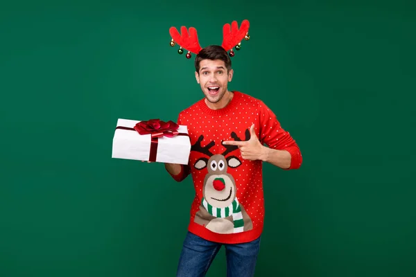 身穿红色针织毛衣的相貌男子指着新年礼物盒孤立的绿色背景 — 图库照片