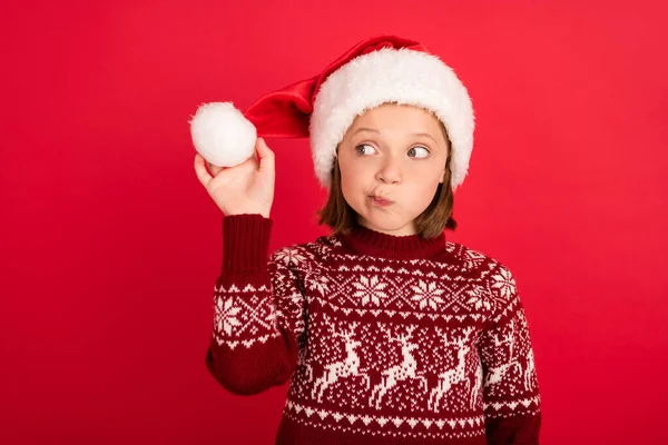 Foto de menina curioso interessado segurar bola fofa desgaste santa chapéu véspera inverno isolado sobre fundo de cor vermelha — Fotografia de Stock