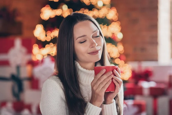 Φωτογραφία από γλυκό γοητευτικό νεαρή γυναίκα ντυμένος λευκό πουλόβερ απολαμβάνοντας ζεστό τσάι κλειστά μάτια χαμογελώντας εσωτερικό δωμάτιο σπίτι — Φωτογραφία Αρχείου