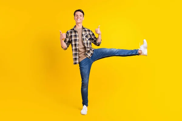 다리를 치켜들고 있는 프로모터 유연 한 사람의 사진, 엄지손가락을 들고 노란색 배경에 고립된 격자무늬 셔츠 청바지를 입고 있는 모습 — 스톡 사진