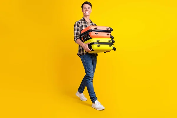 Φωτογραφία του συμπαθητικού άντρα μεταφέρουν stack βαλίτσα με τα πόδια φορούν καρό πουκάμισο τζιν παπούτσια απομονωμένο κίτρινο χρώμα φόντο — Φωτογραφία Αρχείου