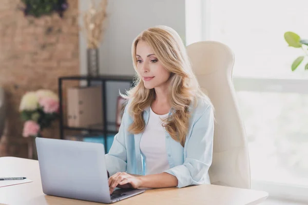 Foto de adorable mujer de edad dulce vestido camisa azul sentado mesa de trabajo moderno gadget sonriendo en el interior de la habitación de casa — Foto de Stock