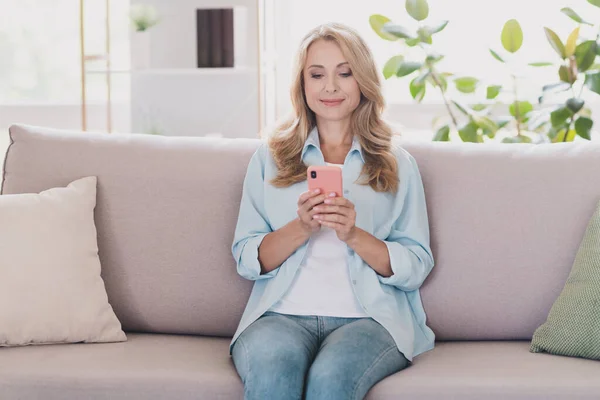 Фотопортрет женщина блондинка волосы улыбается сидя на диване чтение новостей держать смартфон — стоковое фото