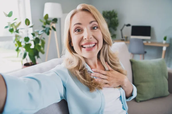 Foto av glad bloggare dam hålla kameran skjuta selfie hand bröst tacksam bära blå skjorta hem lägenhet inomhus — Stockfoto
