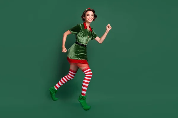 Фото очаровательной милой молодой женщины в костюме эльфа прыгает высоко работает быстро улыбается изолированный зеленый цвет фона — стоковое фото