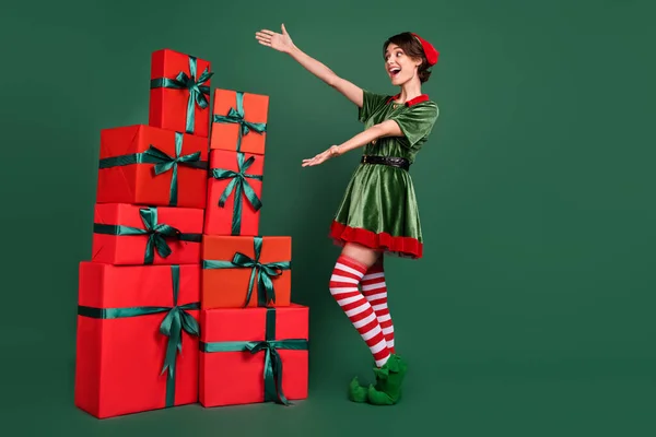 Photo of confident impressed young woman dressed elf costume showing arms gift boxes pile smiling isolated green color background — Stock Photo, Image