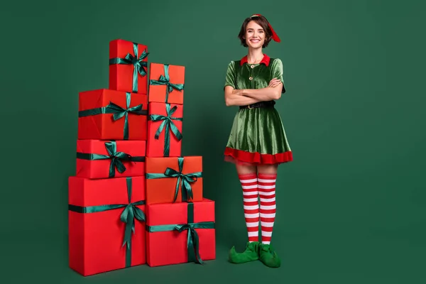 Фото довольно уверенной молодой женщины, одетой в костюм эльфа стоя подарочные коробки скрещенные руки улыбаясь изолированный зеленый цвет фона — стоковое фото