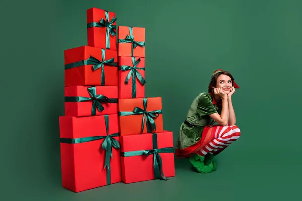 Foto de mulher bonita engraçada vestido traje de elfo sentado perto de caixas de presente pilhas braços bochechas olhando espaço vazio isolado cor verde fundo — Fotografia de Stock