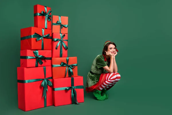 Tatlı, rüya gibi genç bayan elf başlığının fotoğrafı hediyelerin yanında otururken kollarının yanakları izole edilmiş yeşil arka planda gülümsüyor. — Stok fotoğraf