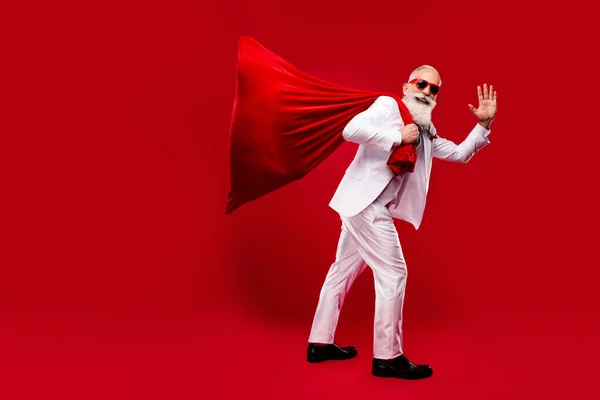 Foto van zorgeloze leuke man gekleed witte kerst kostuum donkere bril met grote zak zwaaiende arm geïsoleerde rode kleur achtergrond — Stockfoto