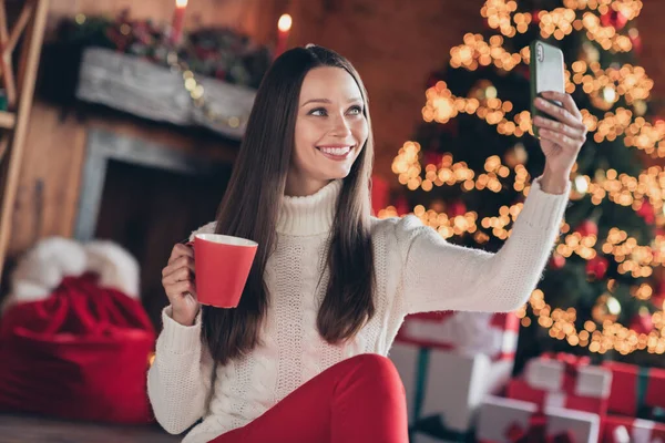 Foto de encantadora joven bonita vestida de punto suéter beber bebida viraje selfie sonriendo en el interior habitación casa —  Fotos de Stock