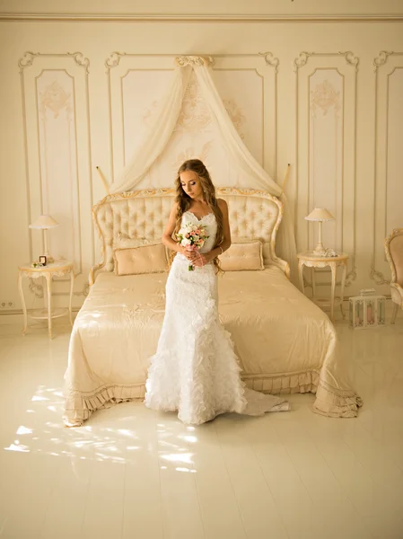 Charming beautiful bride holding wedding bouquet in her hands. bride in white dress in a luxury interior, near the beautiful white bed — Stock Photo, Image