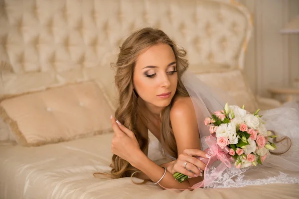 Charmante mooie bruid, bruiloft boeket bedrijf in haar handen. bruid in witte jurk in een luxe interieur liggen op het mooie witte bed — Stockfoto