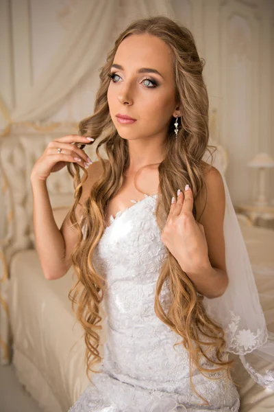Beautiful bride in her bright bedroom sitting on a luxurious white bed and holding her very long hair — Stock Photo, Image