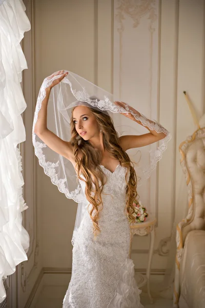 Beautiful bride in her bright bedroom near the window holding her veil — Stock Photo, Image