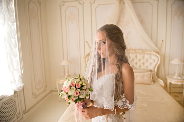 Encantadora novia hermosa celebración de ramo de boda en sus manos. novia en vestido blanco en un interior de lujo, cerca de la hermosa cama blanca. Novia en hermoso velo blanco — Foto de Stock