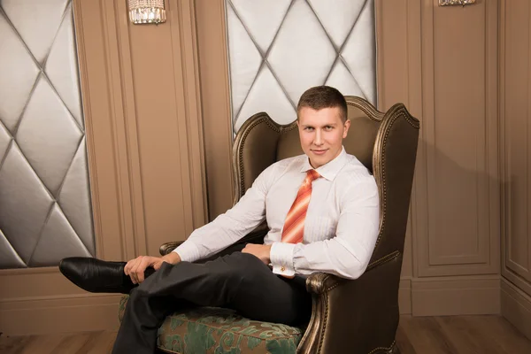 Confidence handsome businessman in chic interior. Successful young man in a business suit sitting on the armchair with his legs crossed and smiling — Stock Photo, Image